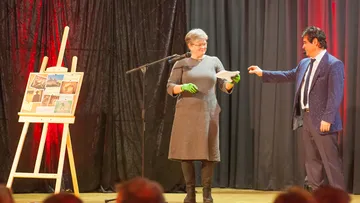 Zwei Personen auf Bühne mit Staffelei, tauschen Objekt.