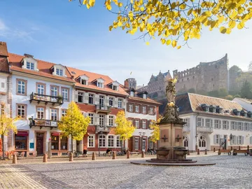 Brunnen in einem Platz, umgeben von historischen Gebäuden und Bäumen.