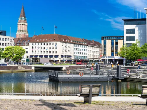 Stadtansicht mit Wasser, Pflanzen, Gebäuden und klarem Himmel.