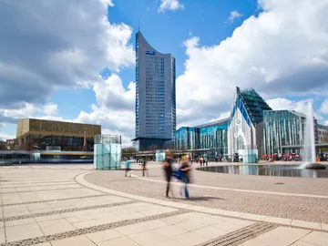 Tall buildings stand in front of a square with people and water.
