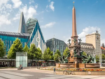 Fountain with an obelisk surrounded by modern buildings and trees.