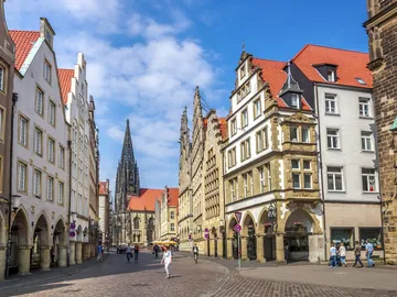 Colourful houses stand, people walk, sunny town square.