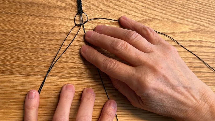 Two hands braiding black thread on wood.