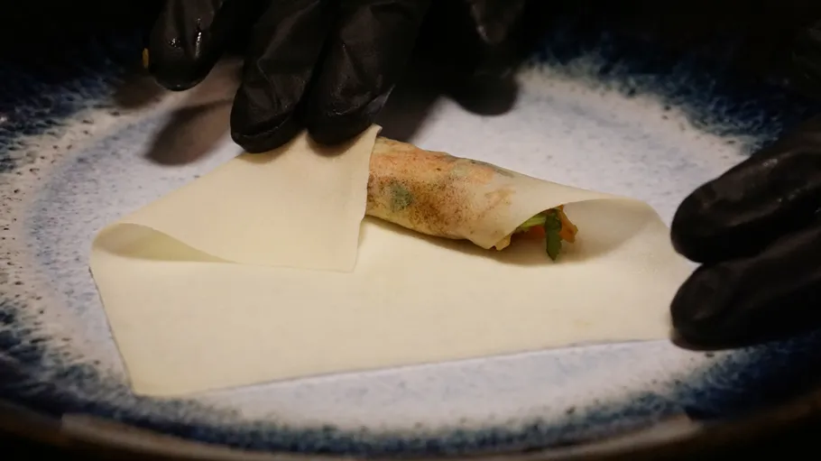Gloved hands rolling spring roll on plate.
