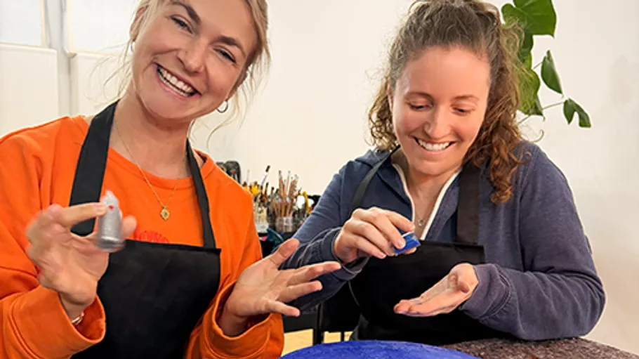 Two women smiling, holding small paint bottles indoors.