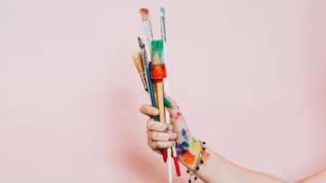 Hand hält bunte Pinsel vor rosa Hintergrund.