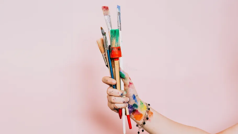 Hand holding colorful paintbrushes against pink background.