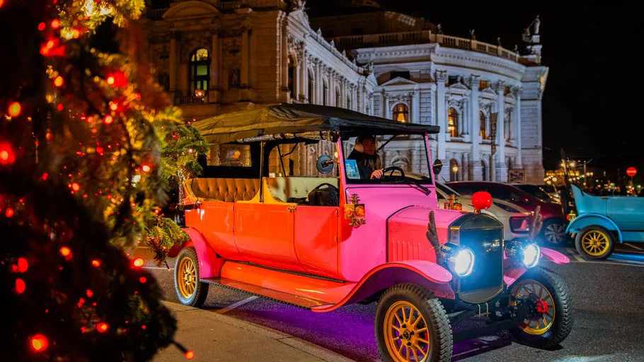 Pink vintage car parked at night, festive lights.