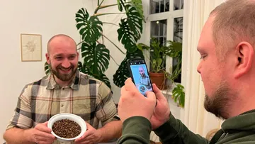 Man holding coffee beans, another takes photo indoors.