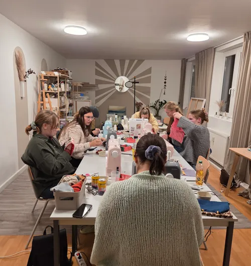 Menschen nähen an einem langen Tisch drinnen.