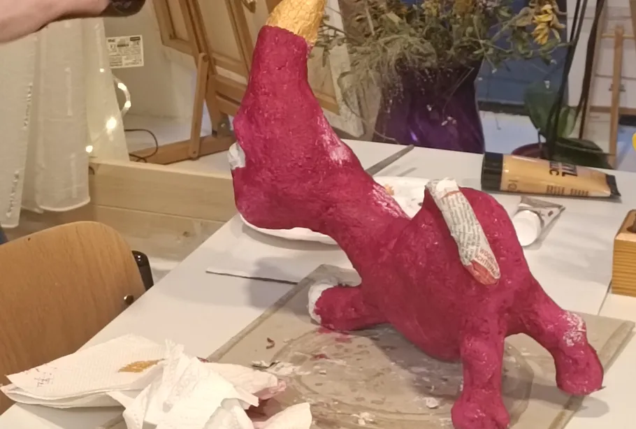 Rosa Skulptur mit Zeitungspapierflügeln auf Tisch.