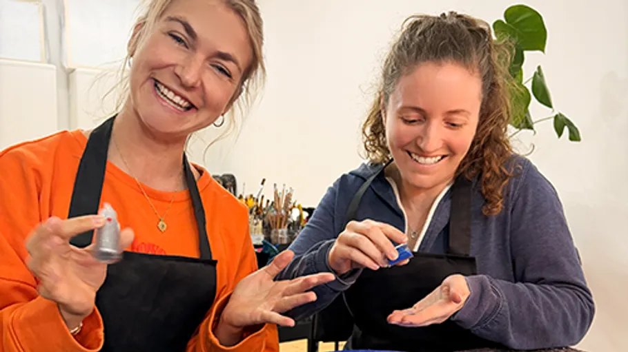 Two women smiling, holding paint bottles indoors.