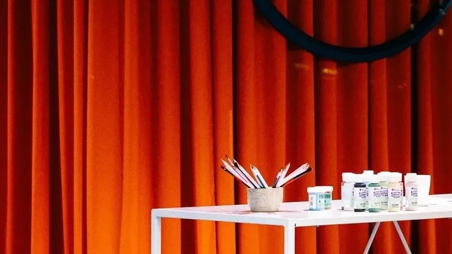 Pencils and paints on table, orange curtain backdrop.