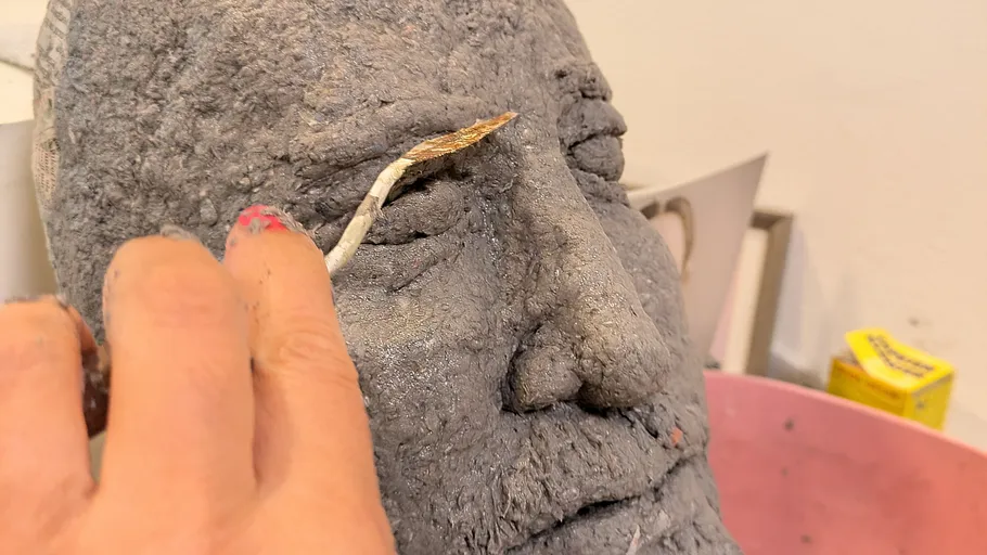 Hand sculpts clay face, indoor work setting.