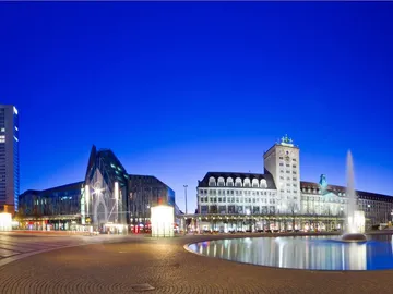 Modernes Gebäude, leuchtet nachts, mit Brunnen in der Mitte.