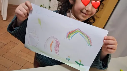 Kind hält Zeichnung mit Regenbogen drinnen.