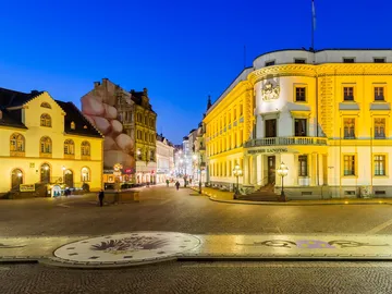 Square with historic buildings, dim lights and pedestrians.