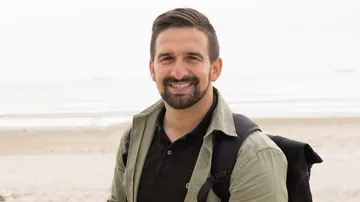 Mann lächelt mit Rucksack am Strand.
