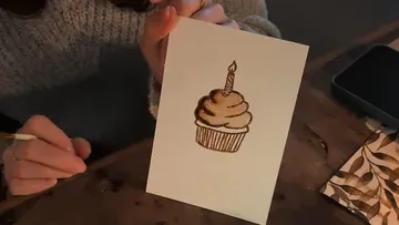 Person zeichnet Cupcake mit Kerze auf Papier.