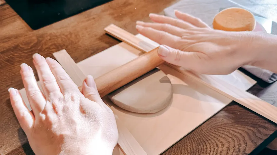 Hands flattening clay with rolling pin on wooden table.