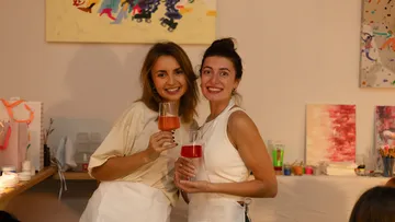Zwei Frauen lächeln, mit Gläsern im Malstudio.