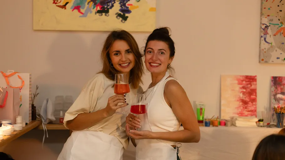 Two women smiling, holding glasses at painting studio.