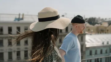 Woman with hat and man outdoors, city background.