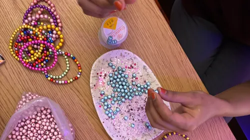 Person making beaded jewelry on wooden table.