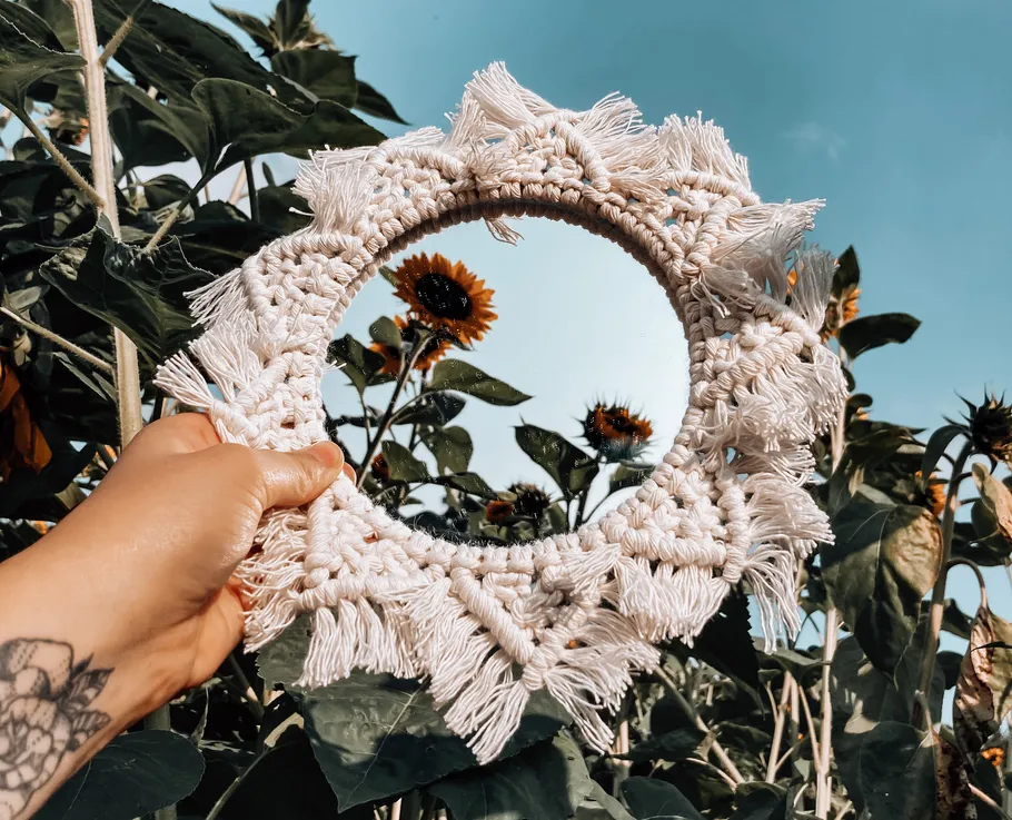 Hand holding macramé mirror in sunflower field.