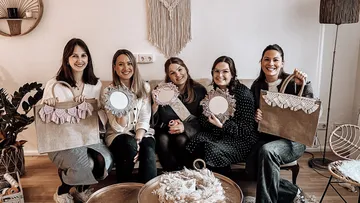 Fünf Frauen sitzen, halten Handwerk, lächelnd drinnen.