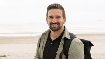 Mann lächelt mit Rucksack am Strand.