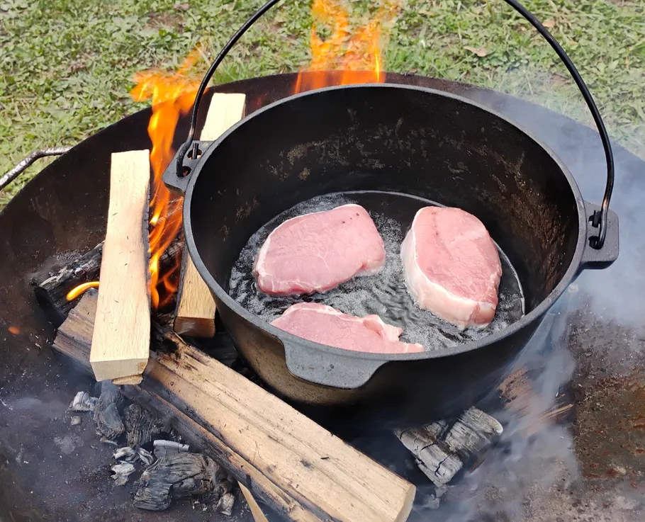 Steaks cooking in pot over open flame.