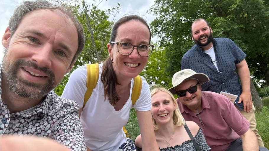 Gruppe von fünf lächelnden Personen im Freien bei Bäumen.