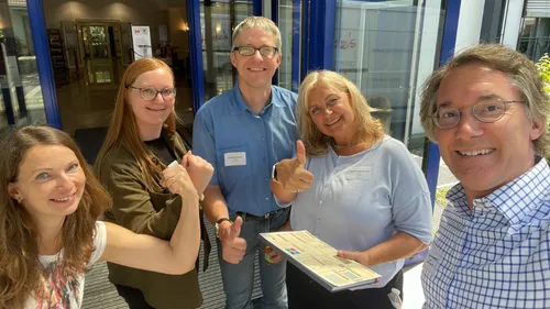 Five people smiling, giving thumbs up indoors.