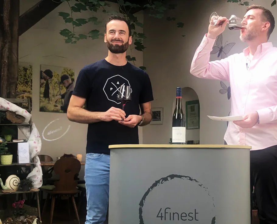 Zwei Männer probieren Wein in Innenraum, Flasche auf Tisch.