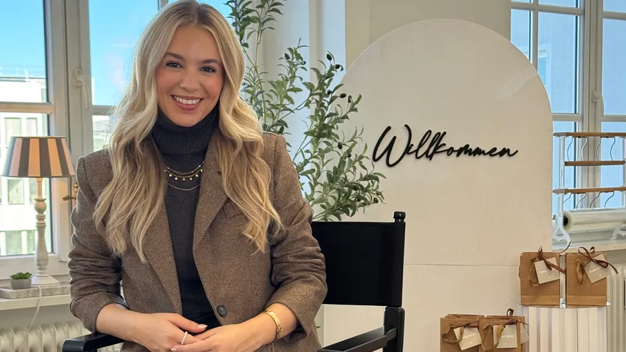 Smiling woman seated, welcome sign in background.