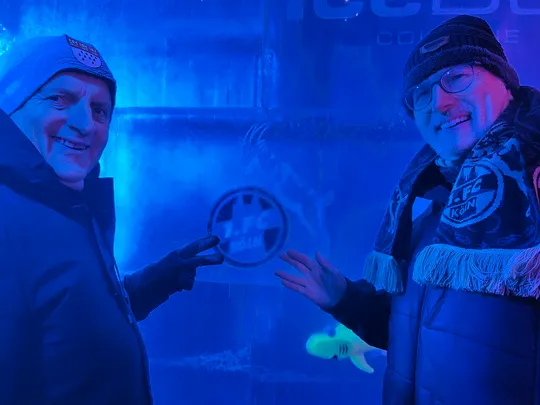 Zwei Männer lächeln neben einer Eiswand.