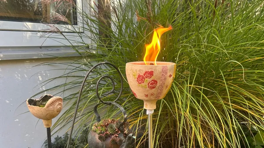 Gartenfackel mit Flamme neben grünen Pflanzen.