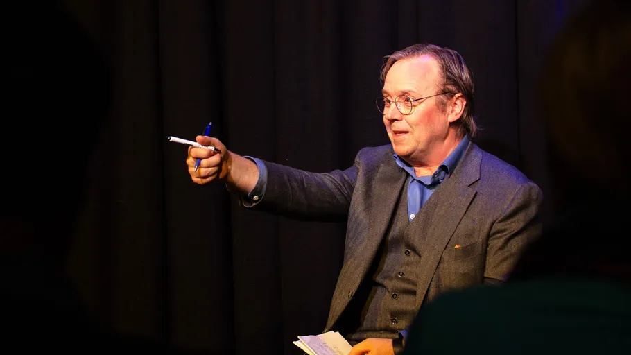 Man gesturing with pen on stage, dark background.