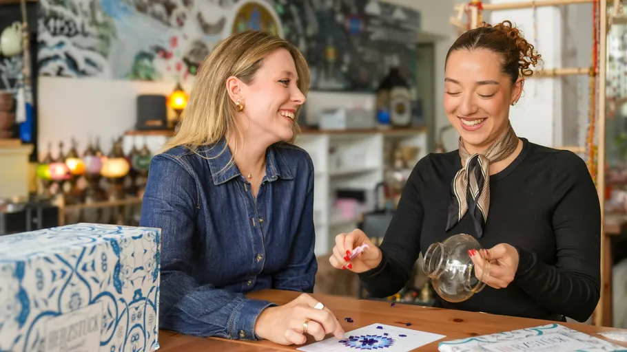 Zwei Frauen lächeln am Mosaiktisch drinnen.