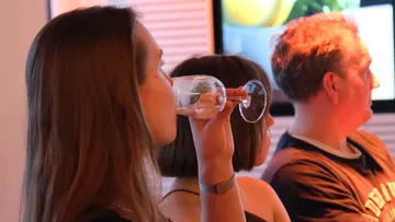People sitting, one drinking wine, in a room.