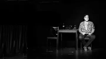 Mann sitzt an Tisch auf Bühne, schwach beleuchtet.