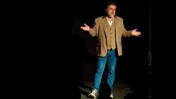 Mann in brauner Jacke gestikuliert auf Bühne.
