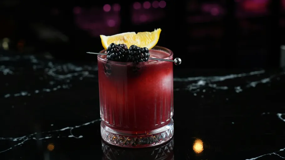 Red cocktail with blackberry and orange garnish.