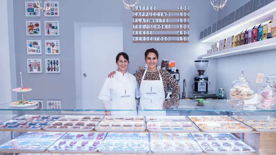 Two women smiling behind cookie shop counter.