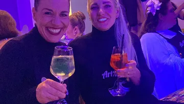 Zwei Frauen lächeln mit Getränken, drinnen.
