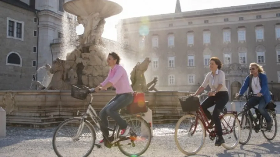 Three people cycling by a fountain in sunlight.