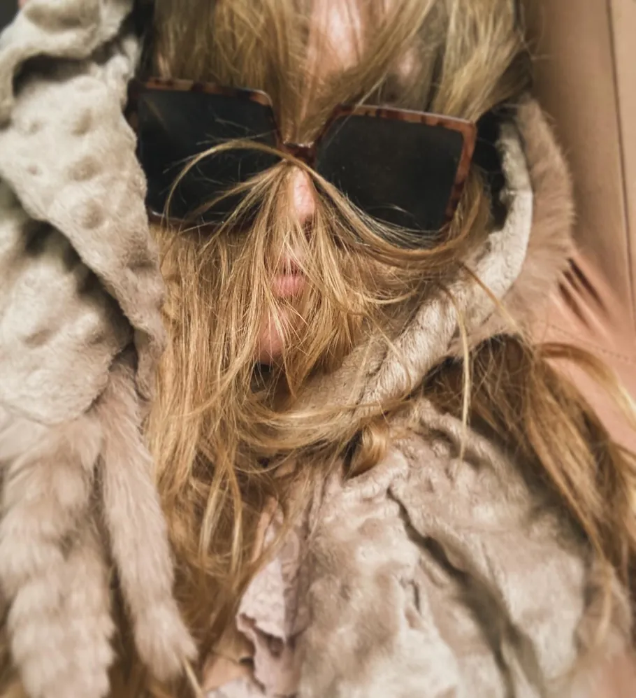 Person with sunglasses, hair covering face, wearing fur.