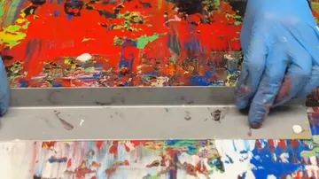 Gloved hands scrape multicolored paint canvas.