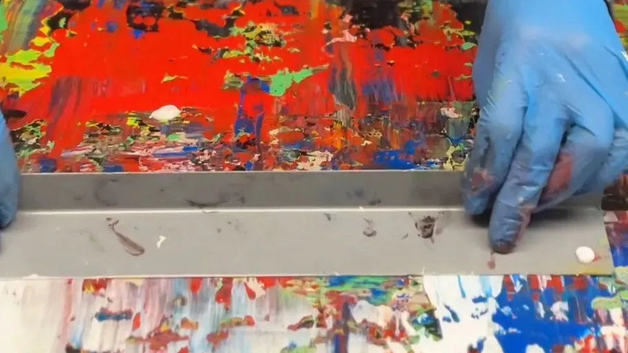 Gloved hands scrape multicolored paint canvas.